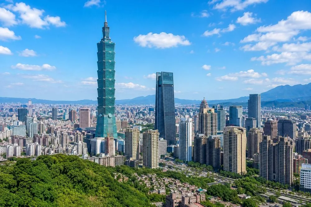 Taipei 101 rises above the Taipei skyline on a sunny day, surrounded by modern skyscrapers, green hills, and distant mountains under a clear blue sky.